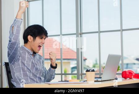 Oui ! Un homme asiatique heureux et enthousiaste se levant le bras pour célébrer le succès. Jeune homme d'affaires utilisant un ordinateur portable à la réception de bureau, il est heureux de le faire Banque D'Images