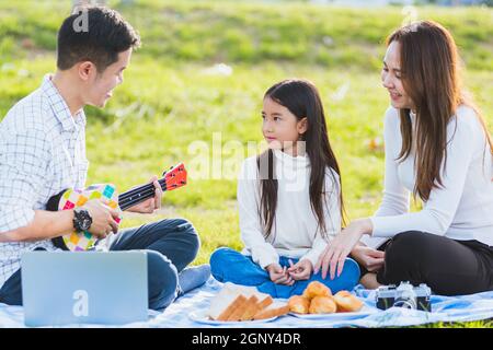 Joyeux père de famille asiatique jeune, mère et enfants s'amuser et profiter de l'extérieur ensemble assis sur la fête de l'herbe avec jouer Ukulele pendant un Banque D'Images