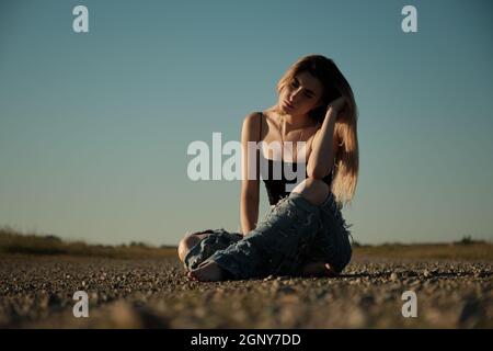 grave triste fille de tramp en jeans déchirés est assis sur une route déserte Banque D'Images