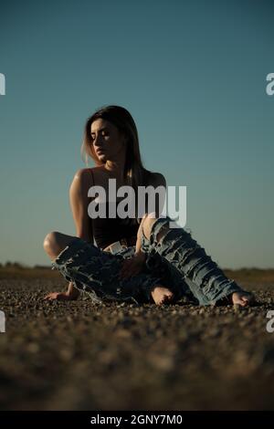 grave triste fille de tramp en jeans déchirés est assis sur une route déserte Banque D'Images