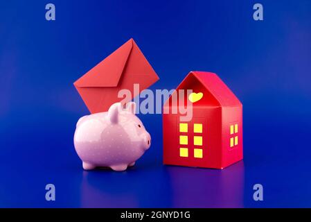 Enveloppe rouge insérée dans une jolie banque de porc rose à côté d'une petite maison rouge de papier avec chaud lumière dans les fenêtres sur fond bleu festif avec Banque D'Images