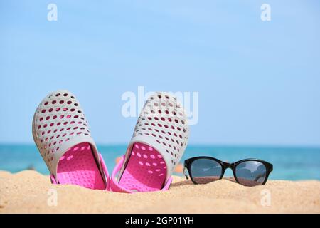 Gros plan sur les chaussures Clogs et les lunettes de soleil noires protectrices sur la plage de sable au bord de la mer tropicale par temps chaud et ensoleillé. Concept de vacances d'été. Banque D'Images