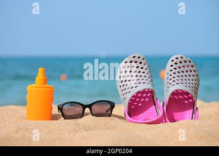 Gros plan sur des chaussures de sabots, de la crème solaire et des lunettes de soleil noires de protection sur une plage de sable au bord de la mer tropicale par temps chaud et ensoleillé. Concept de vacances d'été. Banque D'Images