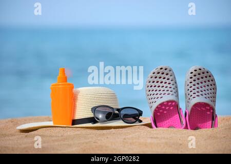 Gros plan sur un chapeau de paille, des chaussures de sabots, de la crème solaire et des lunettes de soleil de protection noires sur une plage de sable au bord de la mer tropicale par beau temps ensoleillé. Vacances d'été con Banque D'Images