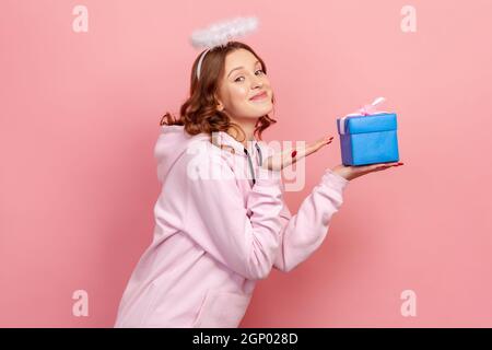 Vue latérale portrait d'une adolescente à cheveux bouclés souriante à capuche et halo sur la tête tenant une boîte cadeau enveloppée de bleu, salutation de vacances. Studio d'intérieur isolé sur fond rose Banque D'Images