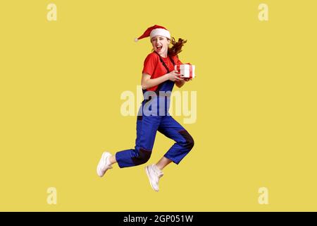 Portrait complet de la femme de livraison sautant haut avec boîte cadeau enveloppée, célébrant Noël, portant une combinaison bleue et un chapeau de père noël. Studio d'intérieur isolé sur fond jaune. Banque D'Images