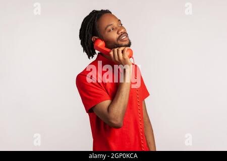 Vue latérale d'un homme avec des dreadlocks et une barbe portant un T-shirt rouge de style décontracté, passer des appels sur un téléphone fixe, regarder l'appareil photo avec un sourire crasseux. Prise de vue en studio isolée sur fond gris. Banque D'Images