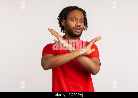 Portrait d'un jeune homme barbu adulte avec des dreadlocks portant un T-shirt rouge, se croisant les mains, avertissement ou interdiction gestuelle, ce qui signifie une finition d'arrêt. Prise de vue en studio isolée sur fond gris. Banque D'Images