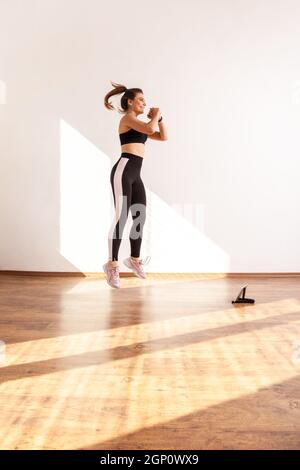 Femme saut, faire des exercices cardio après le blogueur de forme physique, regarder des vidéos tutorial sur tablette, porter des hauts de sport noirs et des collants. Prise de vue en studio pleine longueur éclairée par la lumière du soleil depuis la fenêtre. Banque D'Images