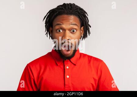 Un homme étonné avec des dreadlocks portant un T-shirt rouge de style décontracté, debout avec la bouche ouverte dans la surprise, a choqué l'expression, entend des nouvelles incroyables. Prise de vue en studio isolée sur fond gris. Banque D'Images