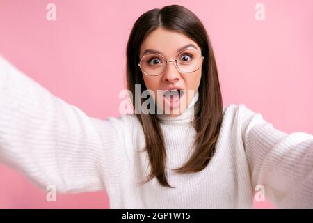 Jeune femme adulte choquée prenant le selfie, ayant un look étonnant et effrayé, point de vue photo, portant blanc style décontracté pull. Studio d'intérieur isolé sur fond rose. Banque D'Images