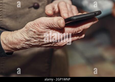 Les mains froissées de la vieille femme tiennent un téléphone mobile moderne. Télécommunications et technologie dans la vie quotidienne. Image à mise au point sélective. Banque D'Images