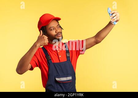 Beau travailleur avec la barbe portant l'uniforme ayant appel vidéo avec le client, montrant le pouce vers le haut, parlant du travail parfait, exécuter l'ordre. Studio d'intérieur isolé sur fond jaune. Banque D'Images