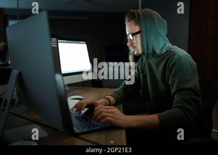Pirate Internet mâle dans la capuche et les lunettes assis aux moniteurs. Programmeur Web illégal sur le lieu de travail, profession criminelle. Piratage de données, cybersécurité Banque D'Images