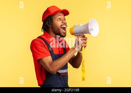 Portrait d'un homme à barbe portant une combinaison bleue et une casquette rouge criant d'annoncer des réductions dans l'industrie des services ou de protester. Studio d'intérieur isolé sur fond jaune. Banque D'Images