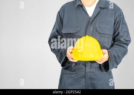 Cloes up Worker debout dans un coverall bleu tenant un casque de sécurité jaune isolé sur fond gris Banque D'Images