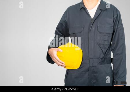 Cloes up Worker debout dans un coverall bleu tenant un casque de sécurité jaune isolé sur fond gris Banque D'Images