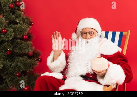 Le Père noël agite la main tout en tenant un cocktail à la noix de coco et assis sur une chaise longue près de l'arbre de noël isolé sur le rouge Banque D'Images