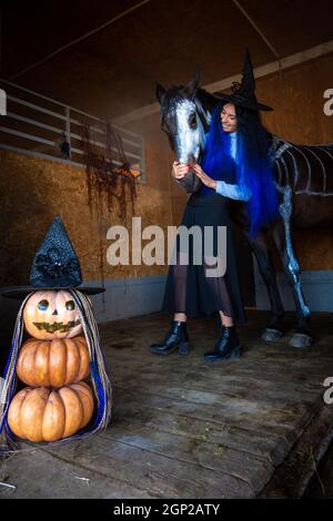 Une fille dans un costume de sorcière embrasse un cheval dans un corral, au premier plan une figure maléfique de citrouilles Banque D'Images