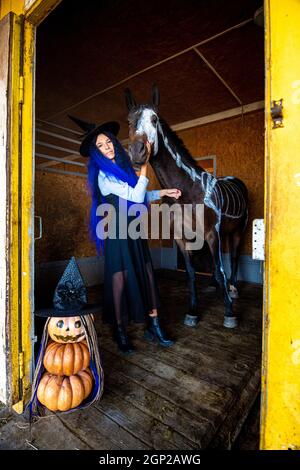 Une fille habillée comme une sorcière se tient dans un corral avec un cheval sur lequel un squelette est peint en peinture blanche, au premier plan est une figurine mal de pompe Banque D'Images