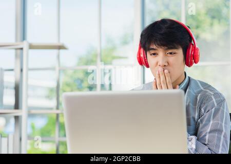 Jeune homme d'affaires asiatique bonheur avec des écouteurs rouges il est assis sur un bureau bureau bureau à la maison à la recherche d'un ordinateur portable moniteur, confiant beau Banque D'Images