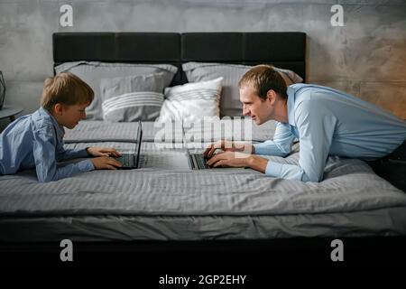 Papa et son fils jouent à des jeux en ligne sur des ordinateurs portables à la maison assis en face l'un de l'autre. La famille passe du temps ensemble le soir près de la maison dans le lit Banque D'Images