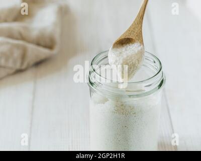 Prêt à faire du pain de pâte de blé de démarrage goutte à goutte à partir d'une cuillère en bois. Bocal en verre avec starter levain une hydratation plus élevée sur fond de bois blanc Banque D'Images