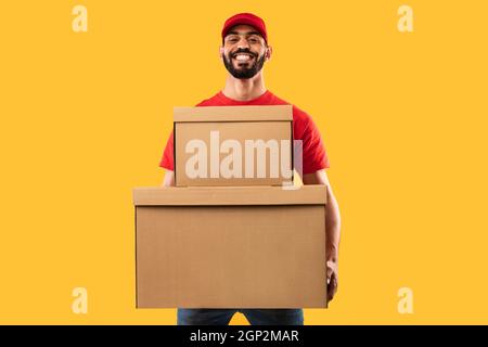 Moyen-Orient Homme Courier tenant de grandes boîtes de colis souriant à la caméra posant en uniforme rouge et en chapeau debout sur fond jaune de studio. Supprimer Banque D'Images