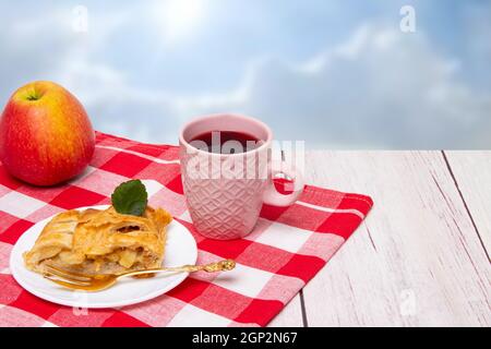 Rouleau de pommes servi. Une tranche de strudel aux pommes fraîches maison avec une tasse de thé et une pomme fraîche sur un tissu de pique-nique rouge sur fond clair. Conce Banque D'Images
