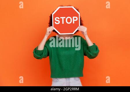 Femme inconnue portant un chandail vert décontracté cachant son visage derrière le symbole rouge d'arrêt de circulation, avertissement, évite les actions interdites. Studio d'intérieur isolé sur fond orange. Banque D'Images