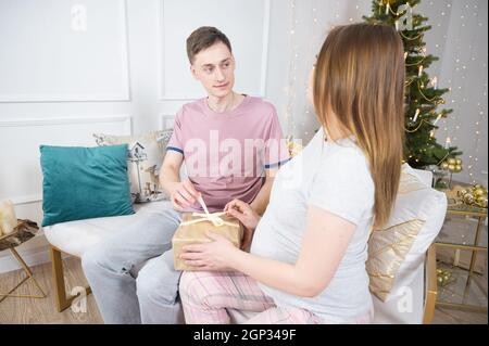 Bonne jeune femme avec mari regardant le cadeau de Noël. Couple échangeant des cadeaux par arbre de Noël Banque D'Images