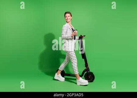 Photo de charmante femme douce habillée de vêtements damés marche kick scooter regardant l'espace vide isolé couleur vert fond Banque D'Images