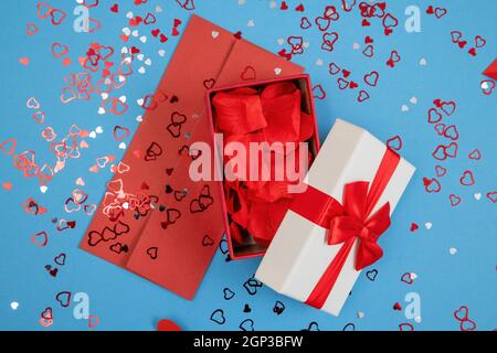 Boîte ouverte remplie de feuilles de rose avec enveloppe rouge sur fond bleu. Cadeau avec carte postale d'amour et coeurs scintillants. Cadeau de l'amour sur le valent Banque D'Images
