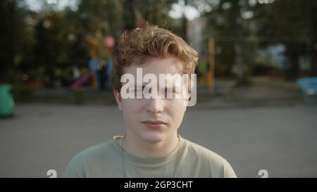 Portrait d'un jeune garçon blond aux yeux, regardant sérieusement vers l'appareil photo à l'extérieur dans un parc verdoyant à l'extérieur Banque D'Images