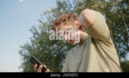 Jeune blonde cheveux coloré eyed garçon luttant pour trouver une réponse à taper un message à son ami ou petite amie sur son smartphone. Belle nature et tre Banque D'Images