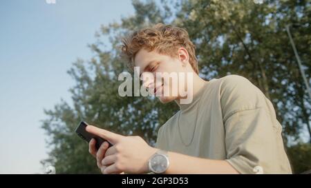 Beau cheveux blond coloré eyed garçon tout en surfant sur l'Internet recevoir de nouveaux messages de notification et de réponses avec un sourire. Belle nature et arbres Banque D'Images