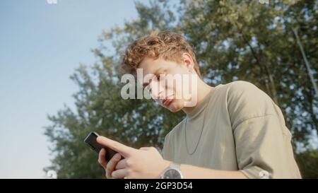 Beau cheveux blond coloré eyed garçon tout en surfant sur l'Internet recevoir de nouveaux messages de notification et de réponses avec un sourire. Belle nature et arbres Banque D'Images