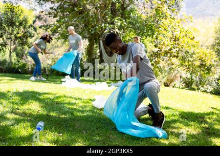 Homme afro-américain souriant tenant un sac de déchets, collectant des déchets plastiques sur le terrain avec des amis Banque D'Images