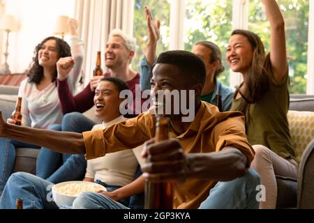 Groupe d'amis féminins et masculins heureux et variés regardant la télévision et buvant de la bière ensemble à la maison Banque D'Images