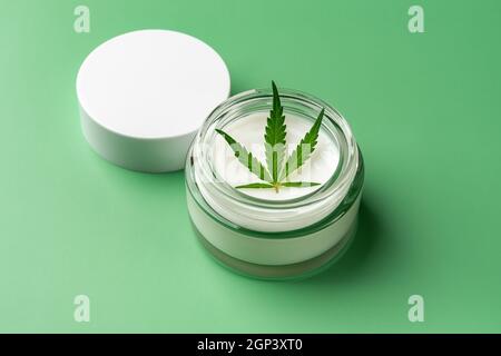 Crème hydratante pour soins de la peau de chanvre et feuille de cannabis fraîche dans un pot en verre ouvert sur fond vert. Cosmétiques à base de plantes de chanvre et usine de marijuana médicale. Banque D'Images