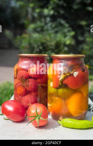 Accueil préservation: Deux grands pots en verre avec des tomates mûres rouges et des poivrons, fermés avec un couvercle en métal, sur une table dans le jardin. Vue avant, cop Banque D'Images