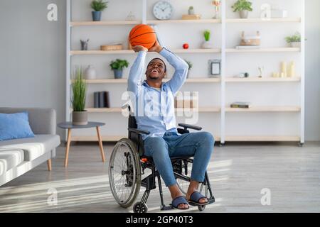 Joyeux homme noir handicapé avec le basket-ball faisant mouvement de lancer, assis en fauteuil roulant à la maison. Jeune homme paraplégique jouant avec le ballon, appréciant le sport Banque D'Images