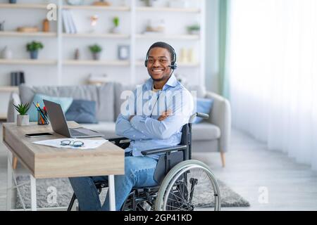 Homme noir paraplégique positif en fauteuil roulant portant un casque, posé devant un ordinateur portable à la maison. Heureux handicapé Afro gars travaillant en ligne dans Banque D'Images