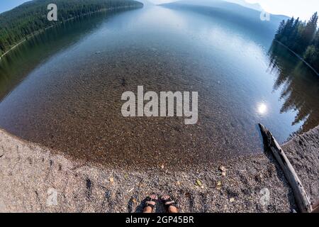 Vue panoramique sur le lac Bowman dans le parc national des Glaciers, avec pieds de femme et sandales sur le rivage Banque D'Images