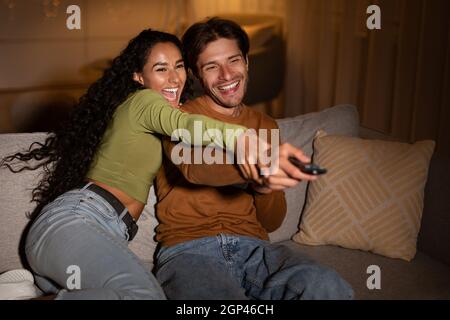 Un couple joyeux de téléspectateurs regardant la télévision pointant la télécommande et les canaux de commutation s'amuser assis à la maison dans la soirée. Week-end détente en famille Banque D'Images