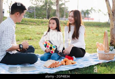 Joyeux père de famille asiatique jeune, mère et enfants s'amuser et profiter de l'extérieur ensemble assis sur la fête de l'herbe avec jouer Ukulele pendant un Banque D'Images