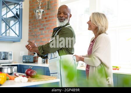 Heureux senior divers couple dans la cuisine de cuisiner ensemble, portant un tablier Banque D'Images