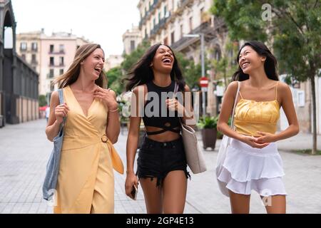 Des jeunes femmes multiraciales riant avec des vêtements d'été élégants, qui profitent de temps libre ensemble tout en marchant dans une rue urbaine Banque D'Images