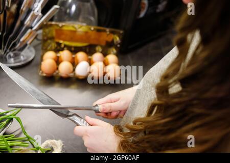 Femme aiguisant couteau de cuisine avec pierre à affûter dans la cuisine, couteau de cuisine. Vue rapprochée des mains. Banque D'Images