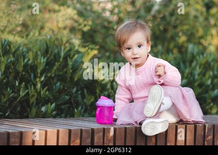 Mignon drôle bébé fille 1 an porter robe rose assis avec bouteille en plastique avec l'eau dans le parc sur fond vert nature gros plan. En regardant le camérier Banque D'Images